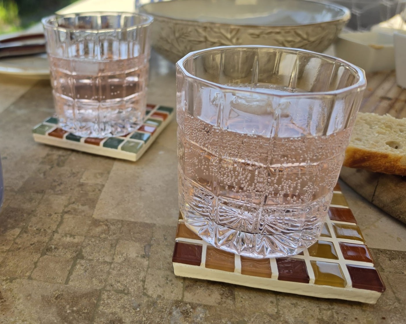 Two mosaic glass coasters on a table in the shade, holding sparkling water