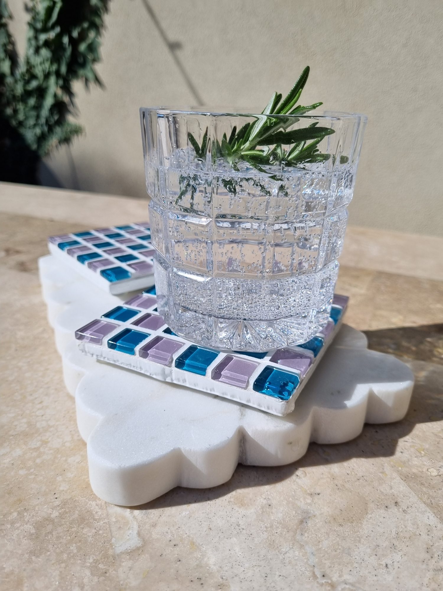 Pigeon Wing handmade glass coaster in teal and lilac grey with a glass of water and rosemary